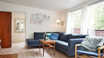 Una sala de estar con un sofá seccional, una mesa de centro de madera y un sillón mecedor de madera. Hay impresiones botánicas enmarcadas en la pared y una ventana con cortinas.