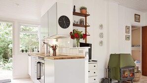 A modern kitchen with a central island, a built-in oven, and a stove. There is a clock on the wall and a window providing a view of greenery outside. - 8 Person Holiday Home in Nexo (Nexø)