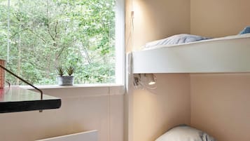Una cama litera con vista a árboles por la ventana, una mesita de noche con una planta, y un aplique en la pared.