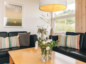 A modern living room with a black leather sofa, a wooden coffee table, and a vase with white flowers. A large window provides natural light, and a framed picture of Denmark is on the wall. - 10 Person Holiday Home in Farso (Farso)