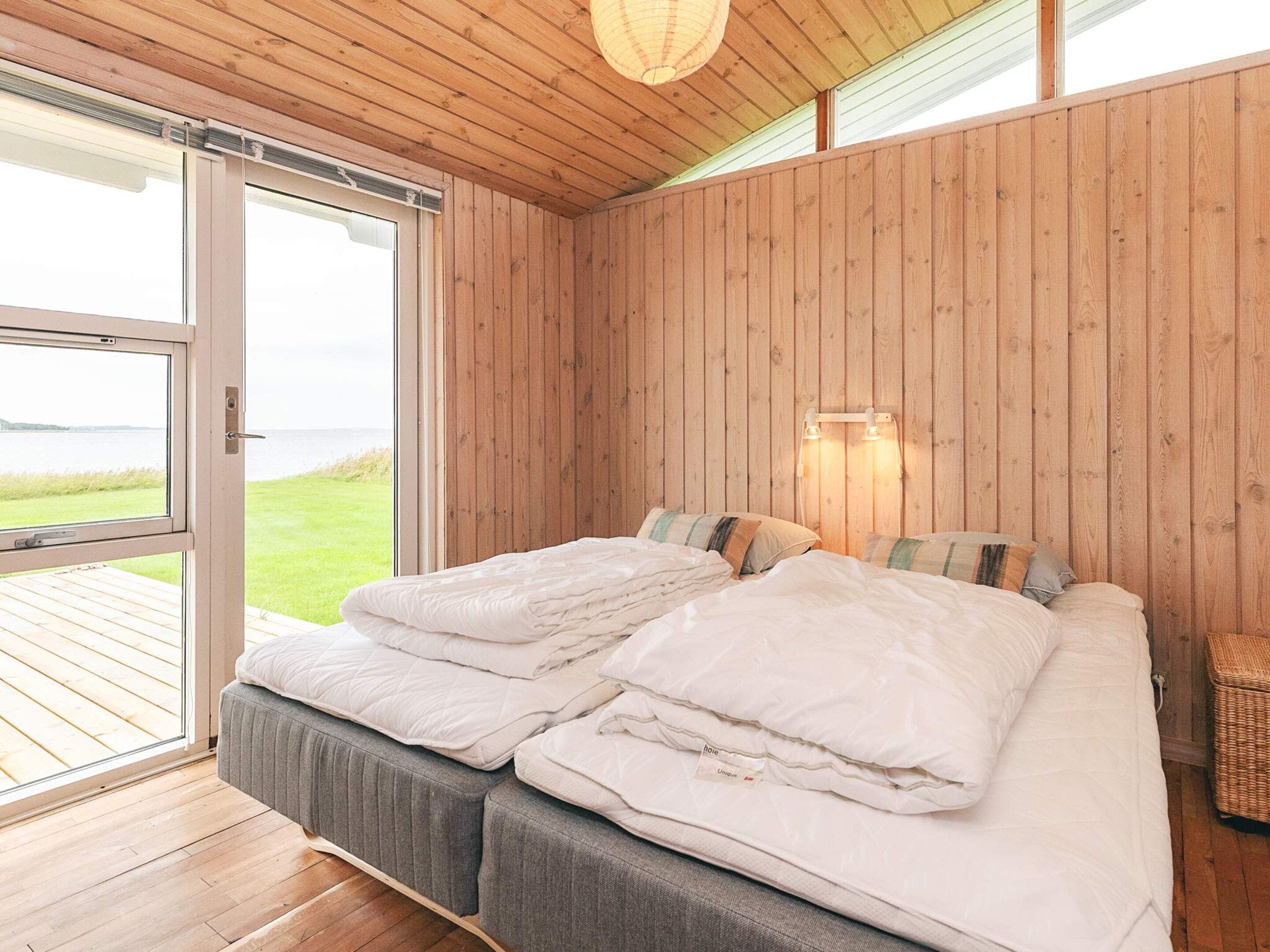 A bedroom with a wooden ceiling, a bed with white bedding, a view of the outdoors, and a wall-mounted light fixture.