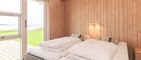A bedroom with a wooden ceiling, a bed with white bedding, a view of the outdoors, and a wall-mounted light fixture.