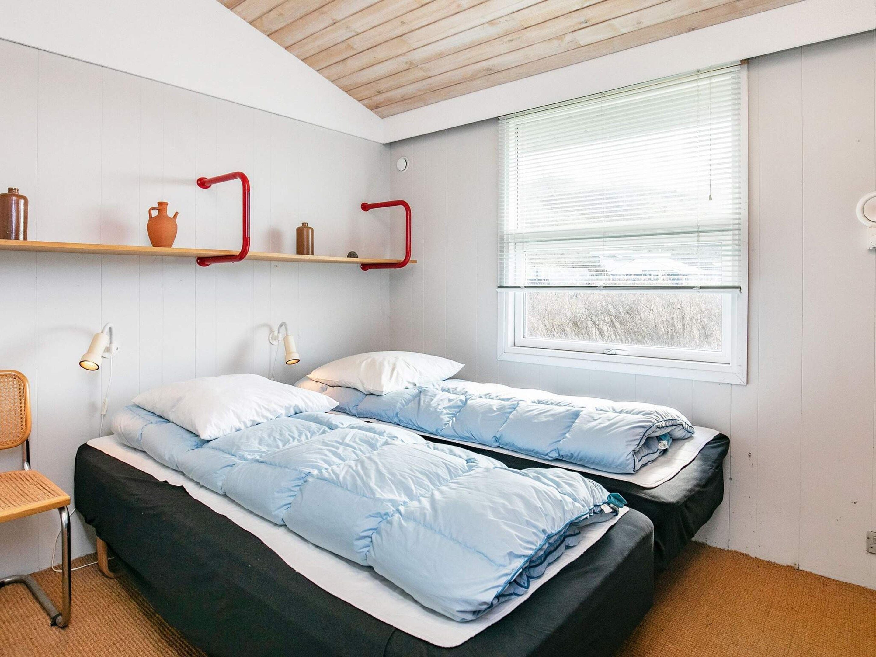 A bedroom with two beds, a window with blinds, a shelf with decorative items, and a chair.