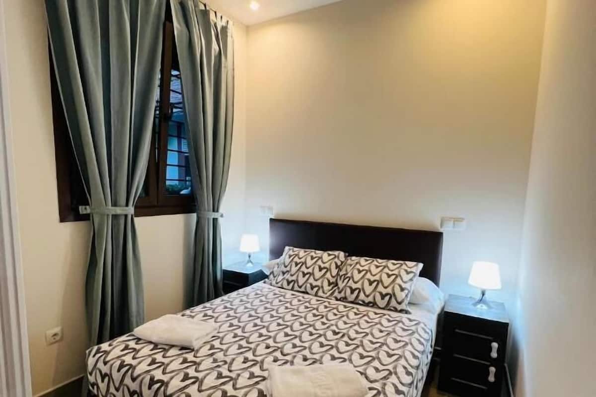 Una cama bien hecha con ropa de cama de estampado de corazones, dos lámparas de noche y una ventana con cortinas.
