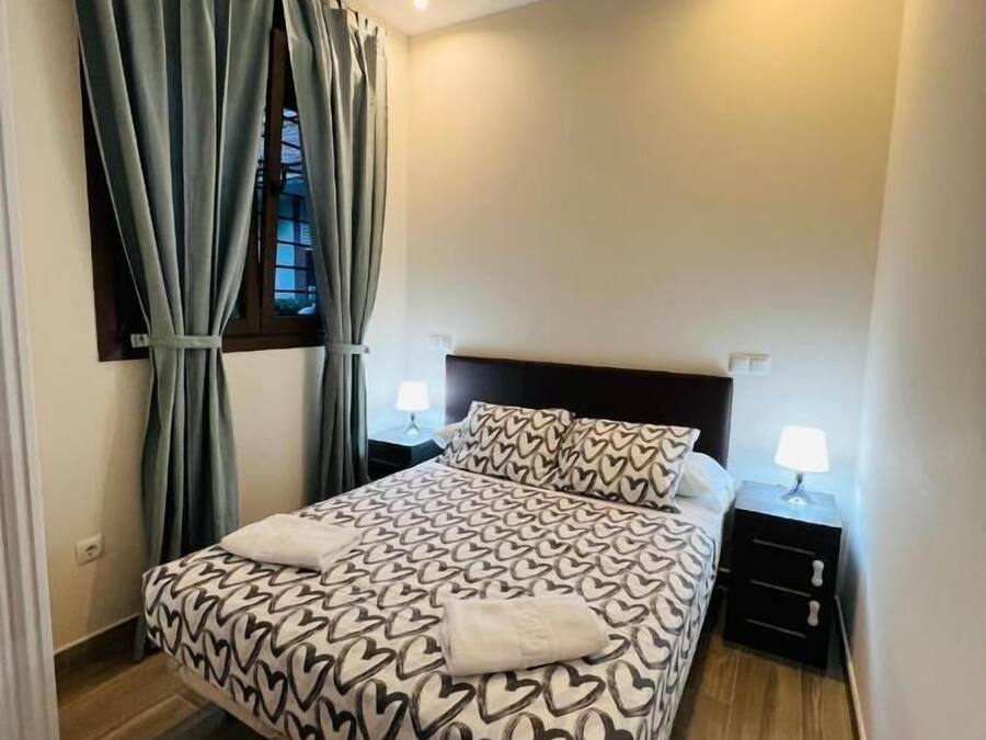 Una cama bien hecha con ropa de cama de estampado de corazones, dos lámparas de noche y una ventana con cortinas.