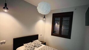 Un dormitorio con cama, mesitas de noche, lámpara colgante, ventana con persianas e iluminación en el techo.