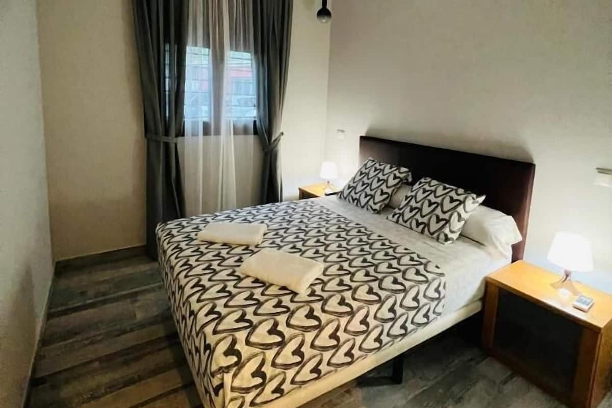 Un dormitorio con cama, mesita de noche, lámpara, ventana con cortinas y luz en el techo.