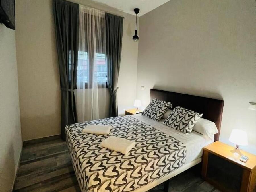 Une chambre à coucher comprenant un lit, une table de chevet, une lampe, une fenêtre avec des rideaux et un luminaire au plafond.