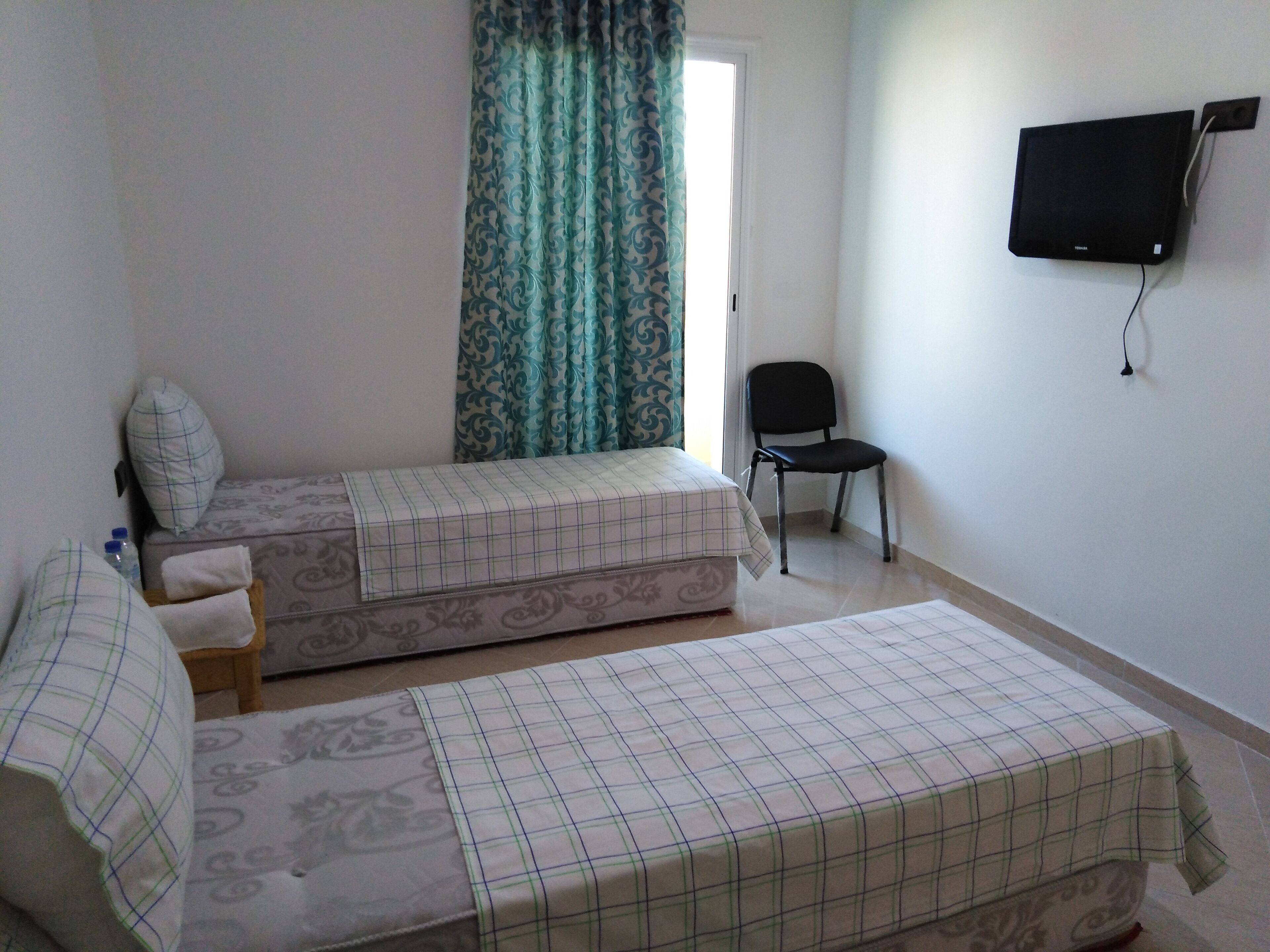 A room with two beds, a chair, a television on the wall, and a patterned curtain.