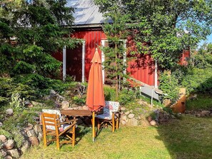 Un bâtiment rouge avec une porte rouge, entouré de verdure et agrémenté d’une table en bois avec des chaises.