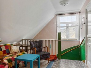 Ett rum med ett sluttande tak, ett fönster med tunna, genomskinliga gardiner, en färgglad soffa, ett blått bord, en svart läderfåtölj och en grön trappa.