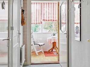 En öppen dörr leder in till ett rum med en stol, ett litet bord och ett fönster med en randig gardin.