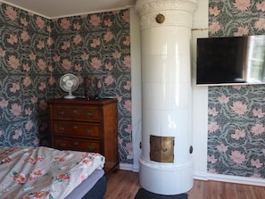 Een slaapkamer met bloemenbehang, een houten dressoir, een bed met bloemenlinnen en een wit, cilindervormig object met een koperen deur, mogelijk een verwarming of opbergunit.