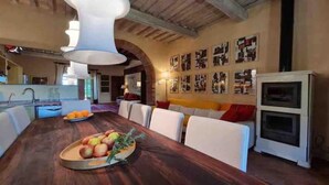 A dining area with a wooden table, white chairs, and a fruit platter. There is a kitchen in the background with a sink and cabinets. - Villa under the Tuscan sun (Toscana)