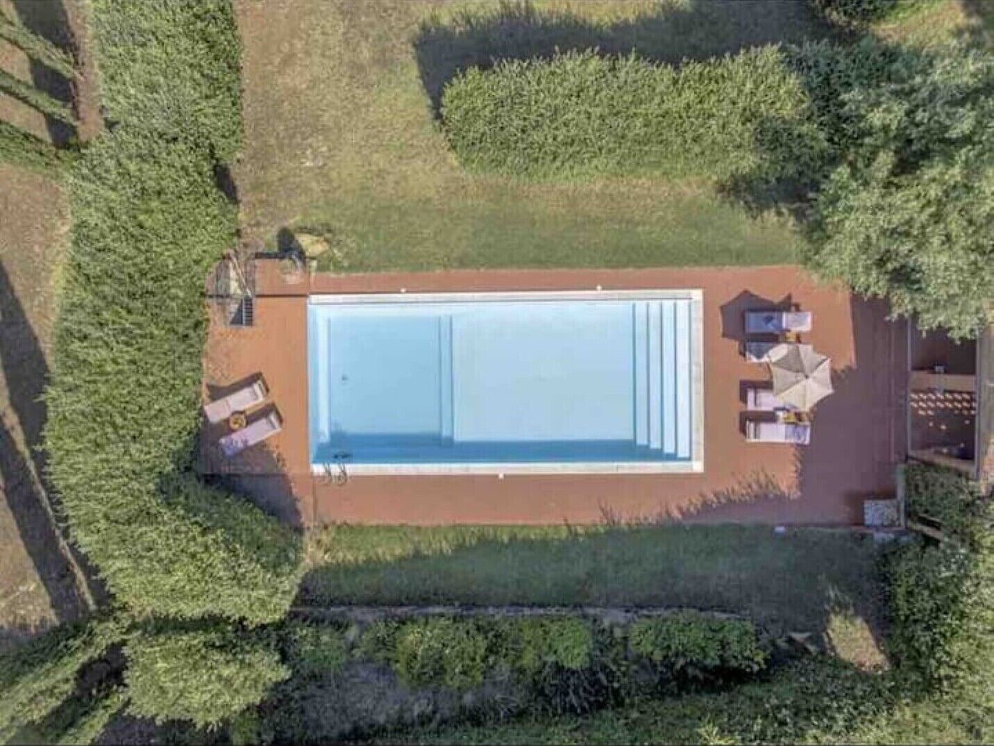 Vista aérea de una piscina rectangular con un solárium, rodeada de vegetación y una valla.