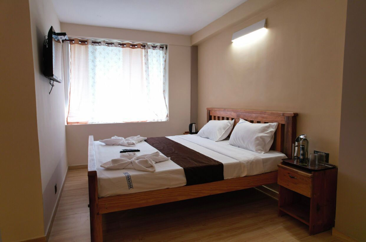 A neatly made bed with a wooden headboard, a bedside table with a lamp, and a window with sheer curtains.