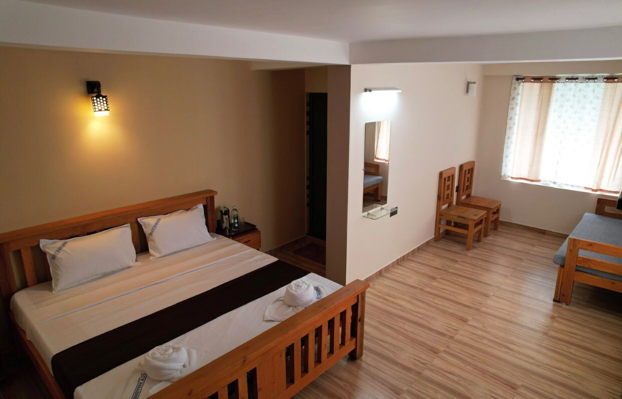 A hotel room with a large bed, a bedside table, a mirror, a small table with chairs, and a window with curtains.