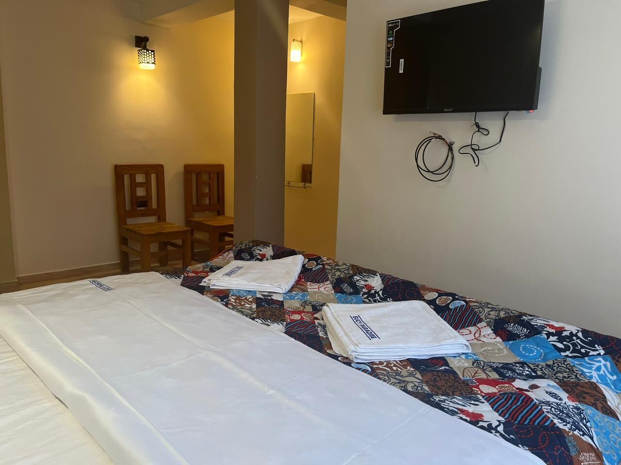 Quarto de hotel com cama, televisão fixada na parede e uma pequena mesa com cadeiras.