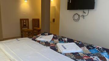 Quarto de hotel com cama, televisão fixada na parede e uma pequena mesa com cadeiras.