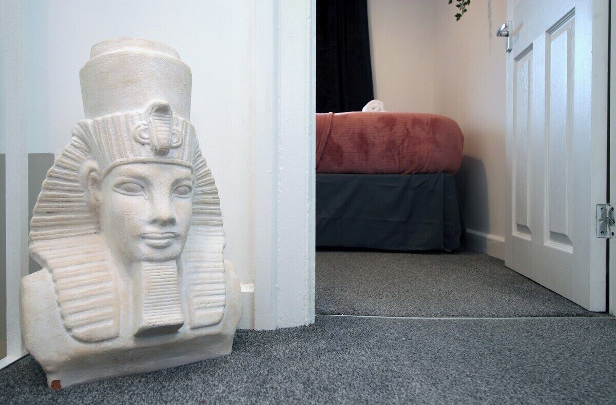 Une statue d’un pharaon se trouve dans un couloir, avec un lit et une porte en arrière-plan.