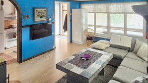 Een woonkamer met een bank, een salontafel en een televisie. In de achtergrond is een keuken te zien.