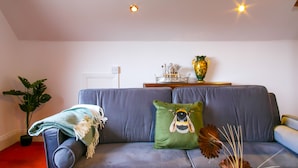 A living room with a grey sofa, a green pillow with a bee design, a potted plant, and a decorative vase on a wooden side table.