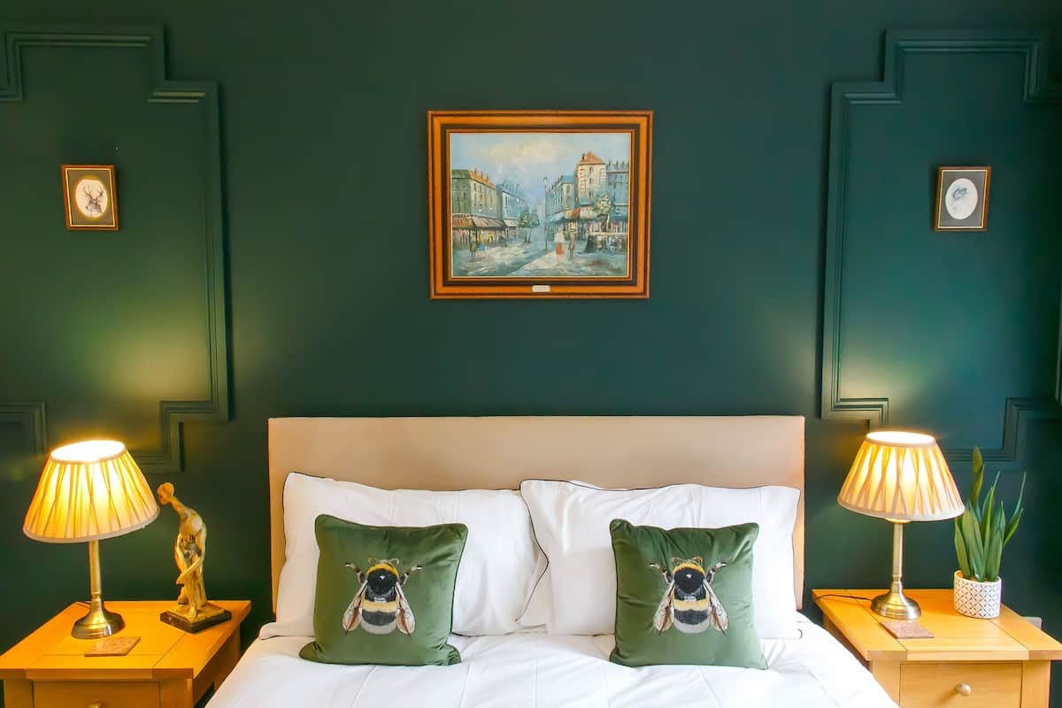 A bedroom with a bed, two nightstands, two lamps, two green pillows with bee designs, a painting of a street scene, and a clock on the wall.