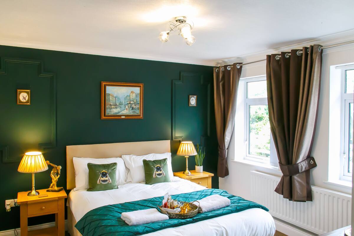 A neatly made bed with a green blanket, two bedside lamps, a framed picture on the wall, and a window with curtains.