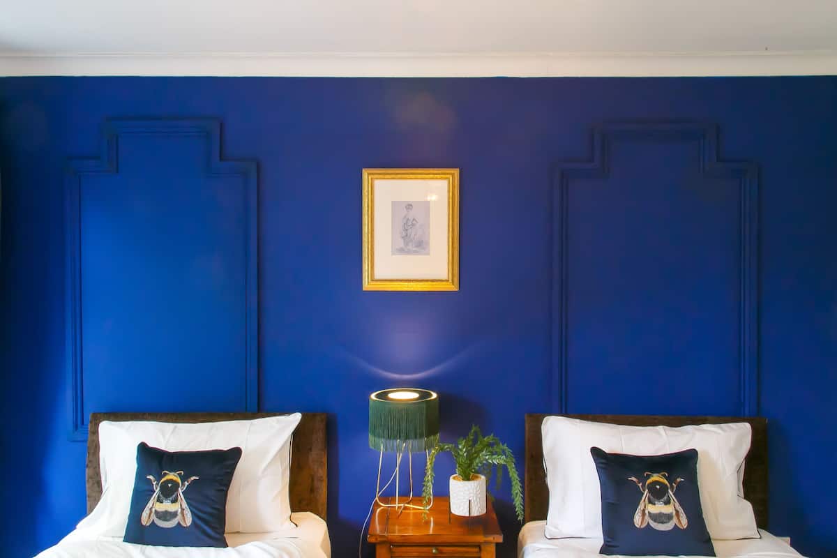 A room with two beds, each with a pillow featuring a bee design, a bedside table with a lamp, and a framed picture on the wall.