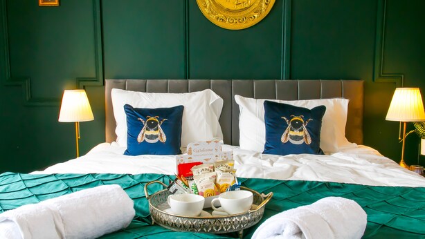 A neatly made bed with a green bedspread, two pillows with bee designs, a tray with cups and a towel, and two wall-mounted lamps.