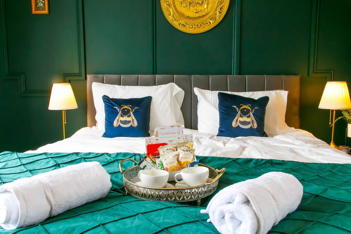 A neatly made bed with a green bedspread, two pillows with bee designs, a tray with cups and a towel, and two wall-mounted lamps.