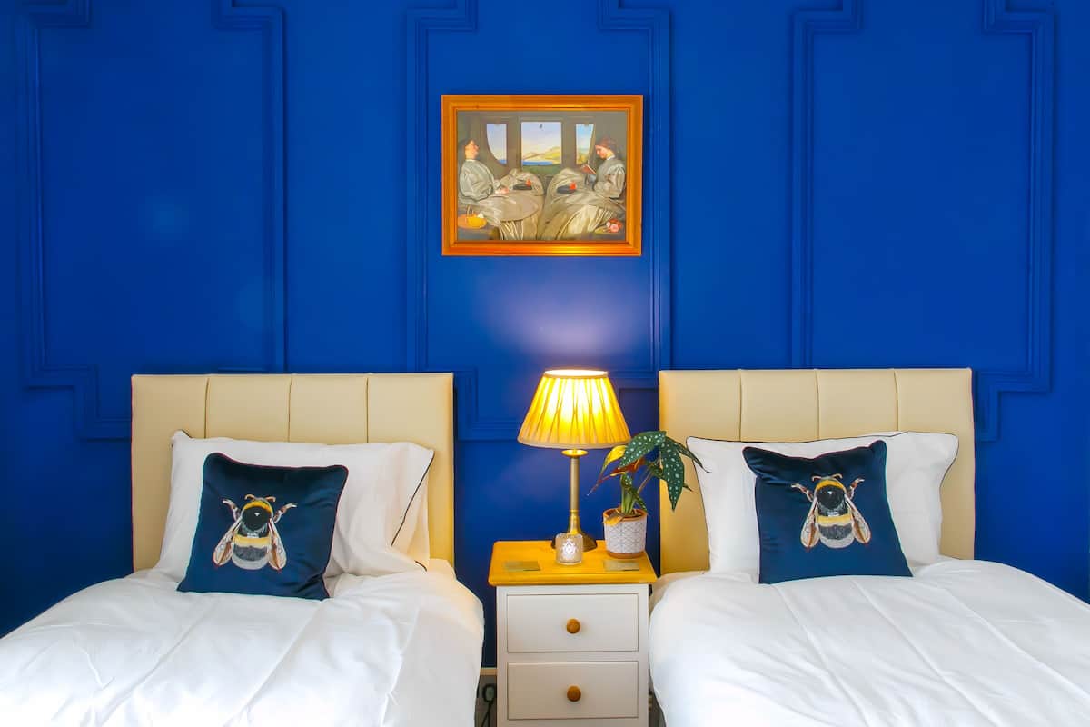 A hotel room with two beds, each with a bee-themed pillow, a bedside lamp, and a framed painting above the beds.