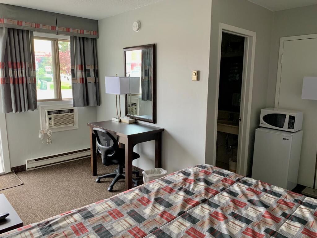 A hotel room with a bed, a desk with a chair, a mirror, a window with curtains, an air conditioner, a microwave, and a door.
