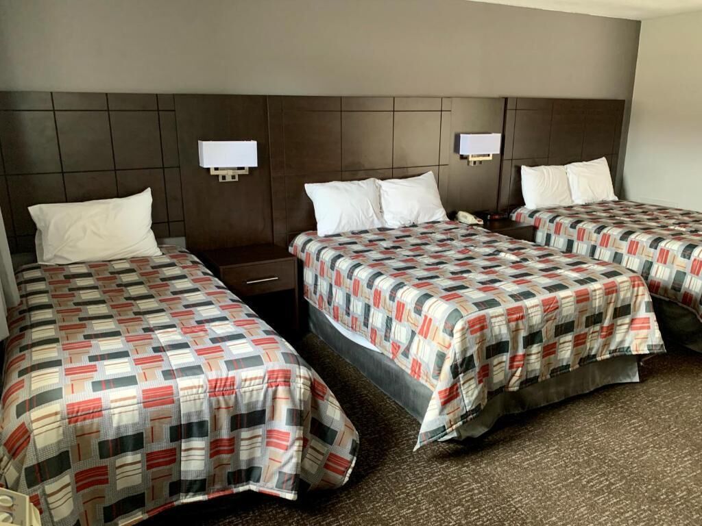 Ein Hotelzimmer mit drei Betten, jedes mit einer gemusterten Tagesdecke, einem Nachttisch und einer an der Wand befestigten Lampe dazwischen.