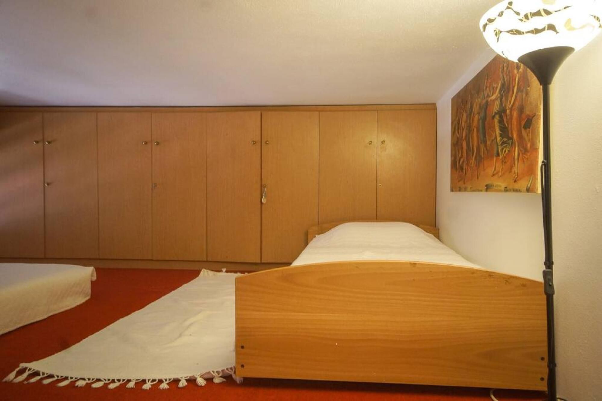Un dormitori amb un llit de fusta, un edredó blanc, una catifes vermella, un armari de fusta i un quadre a la paret.