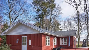 A red wooden building with a white door and windows, a patio with outdoor furniture, and a stone structure in the background. - 4 Person Holiday Home in Ronne-by Traum (Ronne)