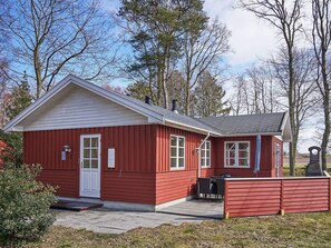 A red wooden building with a white door and windows, a patio with outdoor furniture, and a stone structure in the background. - 4 Star Holiday Home in Ronne (Ronne)