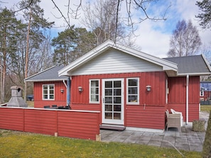 A red wooden cabin with a white door and windows, a chimney, and a small patio area with chairs.