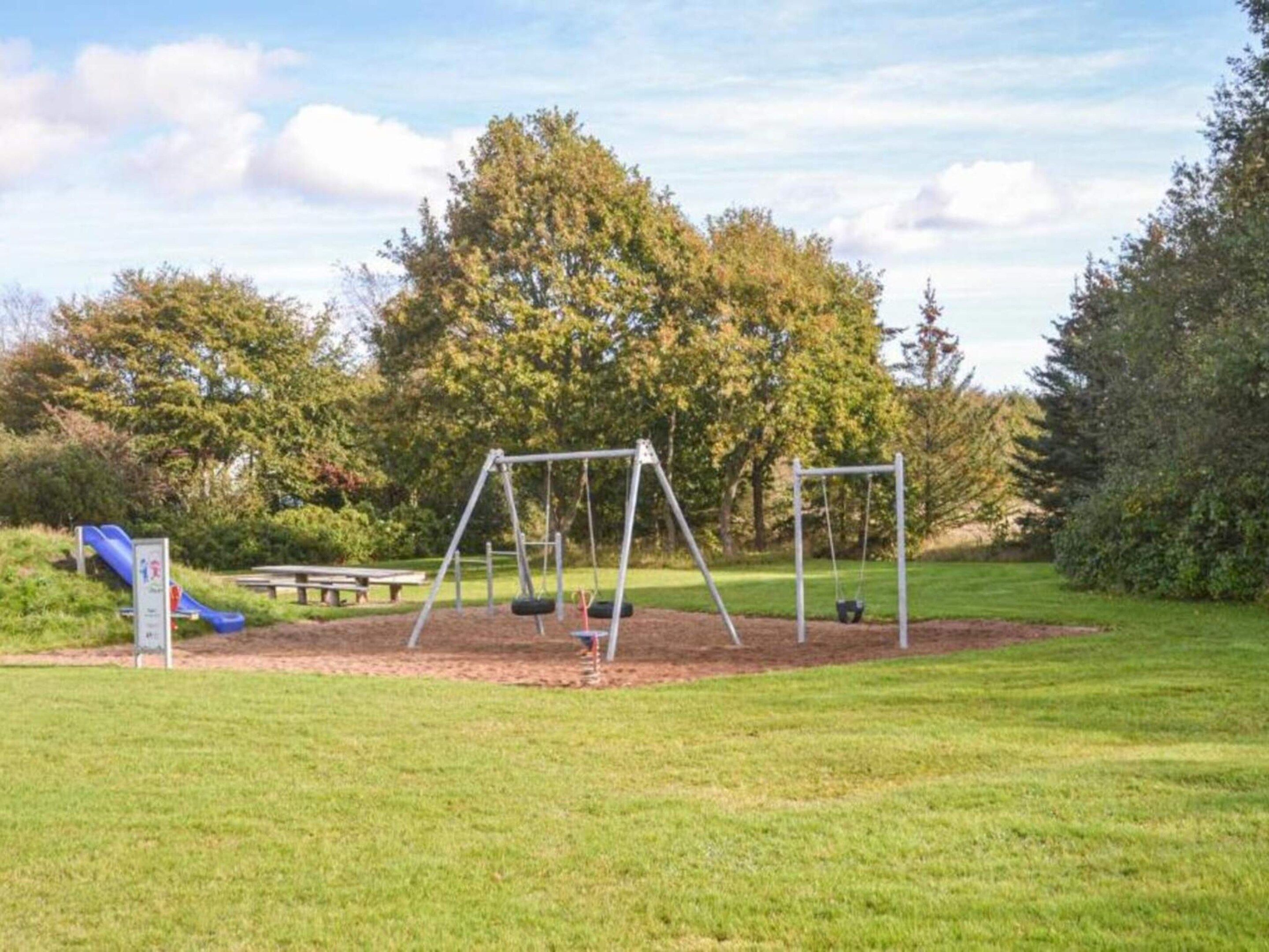 Un parque infantil con columpios y un tobogán, rodeado de césped y árboles.