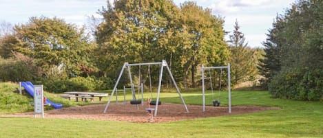 A playground with swings and a slide, surrounded by grass and trees.