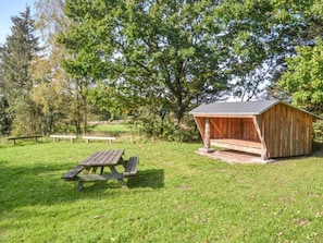 A rustic wooden shed with a sloped roof, a picnic table, and benches in a grassy area with trees in the background. - 5 Person Holiday Home in Hovborg (Hovborg)