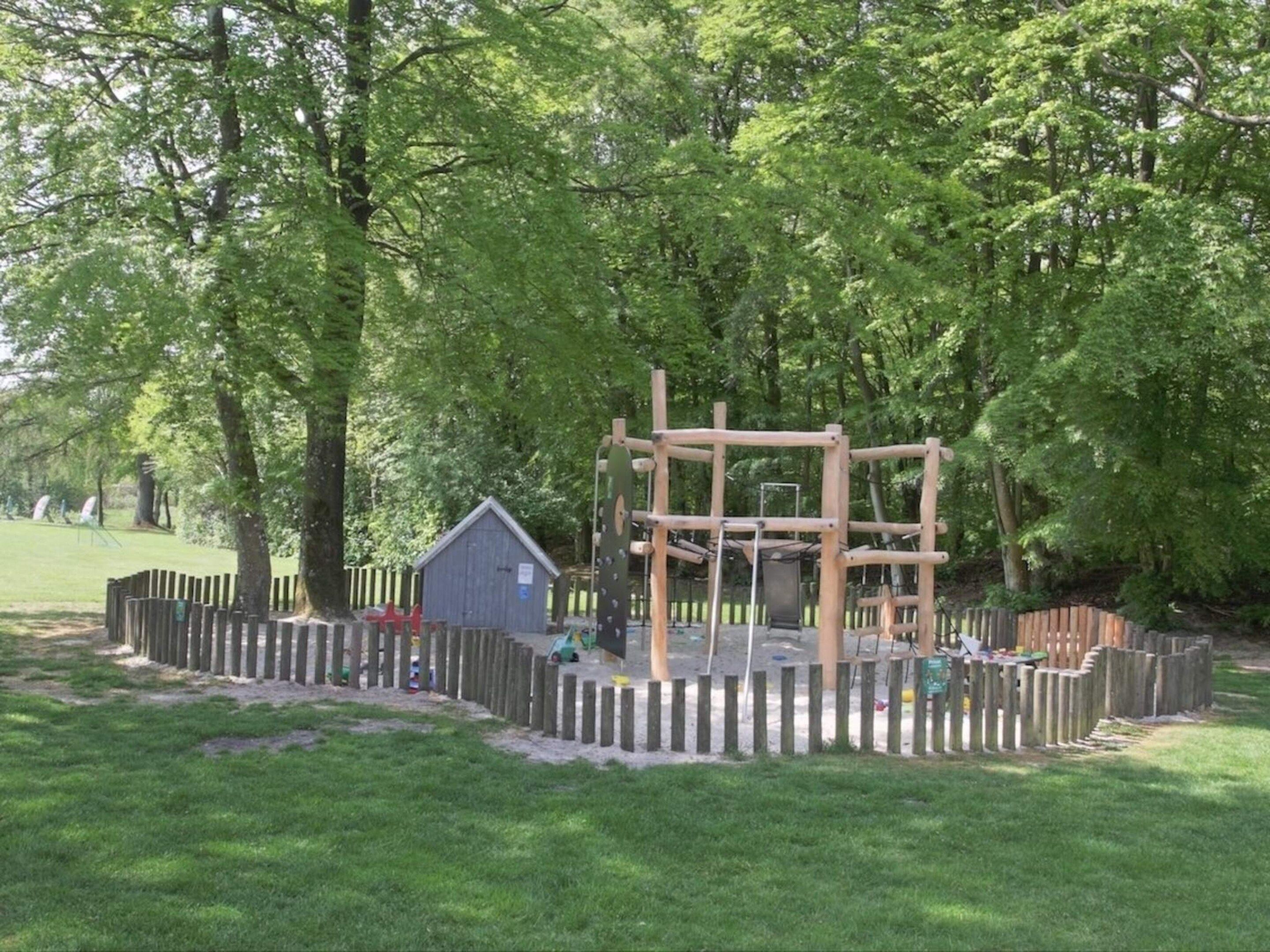 Une aire de jeux avec une structure d’escalade en bois, un petit abri et une zone délimitée par une clôture.