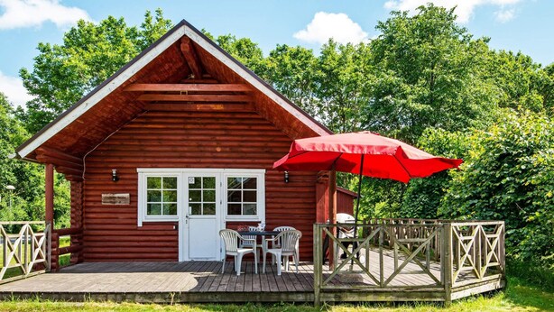 A wooden cabin with a deck, a table, and chairs, surrounded by greenery. - 5 Person Holiday Home in Hovborg (Hovborg)