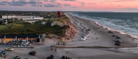 A coastal road with parked cars, a beach, and a building with a red structure on a hill.