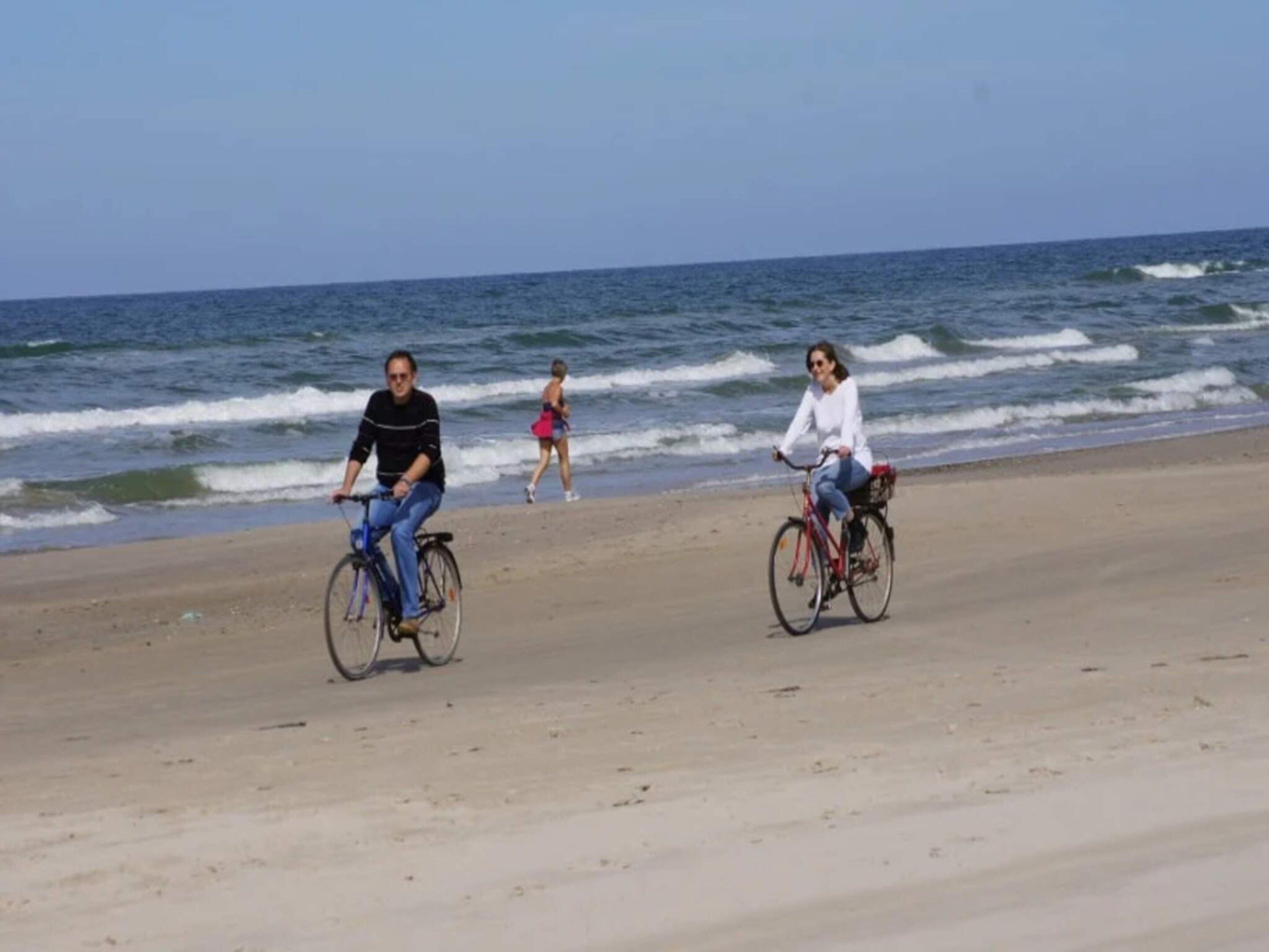 Two individuals riding bicycles on a sandy beach with the ocean in the background.