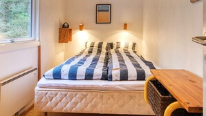 A bedroom with a bed, a wooden table, a wall-mounted clock, and a window with a view of trees.