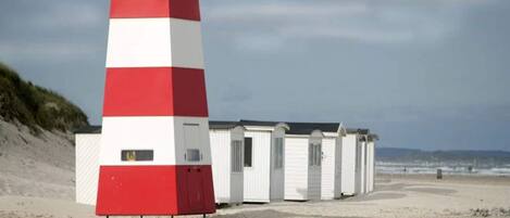 Un faro rojo y blanco al lado de una fila de cabañas blancas en la playa, con una playa de arena y un cielo nublado al fondo.