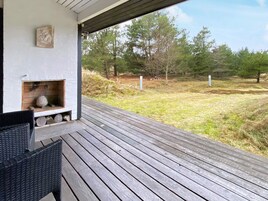 Una terraza de madera con chimenea, mobiliario de mimbre y vista a un área verde con árboles.