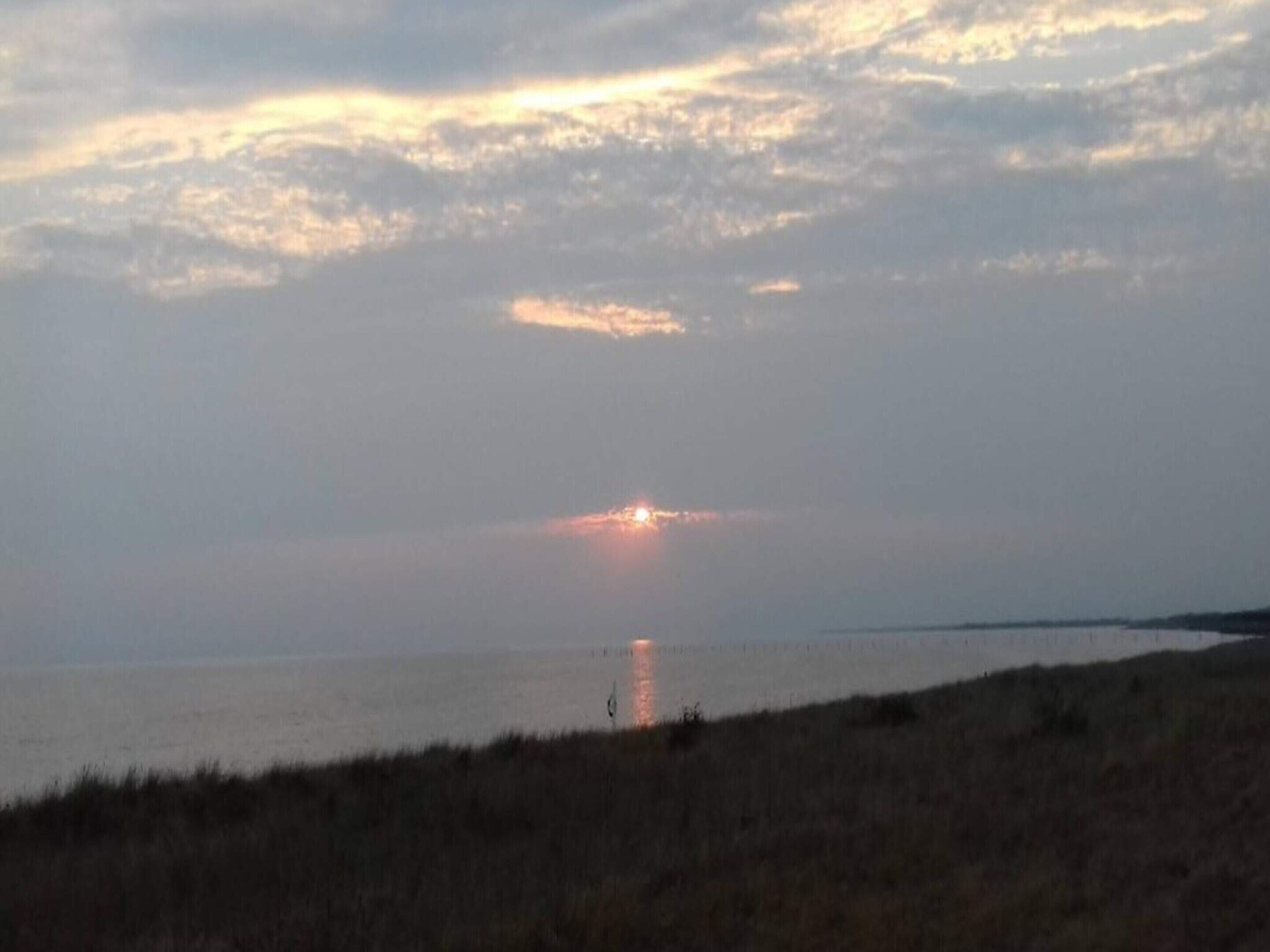 Solnedgang over vand med en fjern kystlinje og en græsbevokset forgrund.