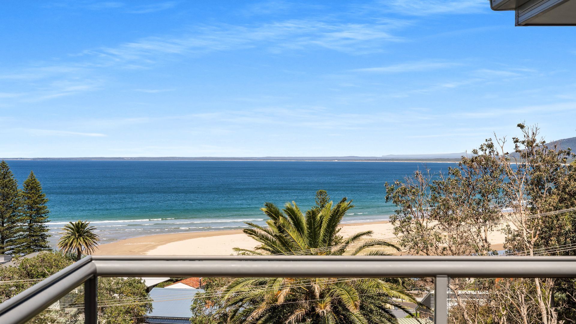 A coastal view from a balcony with a clear sky, a sandy beach, and palm trees.